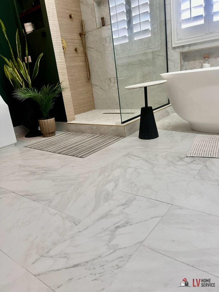 A modern bathroom with sleek white marble tiles, a freestanding bathtub, glass shower, potted plant, and a small black side table